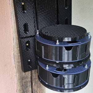 Peut inclure: Un support mural noir imprimé en 3D. Il comporte un panneau arrière texturé et deux compartiments circulaires à plusieurs niveaux avec des accents bleus. L'unité est fixée à un poteau en bois.