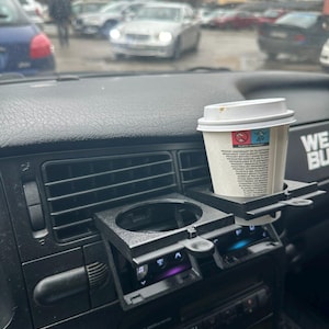 Puede incluir: Un portavasos de coche negro con un vaso de café blanco en su interior. El portavasos está sujeto al salpicadero del coche. El vaso de café tiene una tapa negra y una etiqueta blanca con texto en un idioma extranjero.