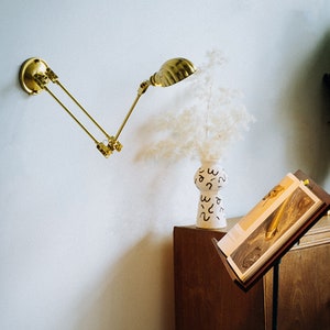 K&ouml;nnte beinhalten: Eine Wandlampe aus Messing mit verstellbarem Arm und gew&ouml;lbtem Schirm. Die Lampe befindet sich &uuml;ber einem Holzschrank, neben einer wei&szlig;en Vase mit schwarzem abstraktem Design und einem Buchst&auml;nder mit einem Buch.