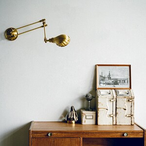 K&ouml;nnte beinhalten: Eine Wandlampe aus Messing mit verstellbarem Arm und muschelf&ouml;rmigem Schirm. Die Lampe ist an einer wei&szlig;en Wand &uuml;ber einem Holzschrank mit dekorativen Gegenst&auml;nden befestigt, darunter ein gerahmtes Bild.