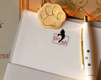Puede incluir: Un clip de papel de pata de gato de oro con una silueta de gato negro y el texto "Chat Noir" en rojo. El clip de papel está en una hoja de papel blanca con un bolígrafo.