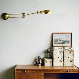 K&ouml;nnte beinhalten: Eine Wandleuchte aus Messing mit verstellbarem Arm ist an einer wei&szlig;en Wand befestigt. Darunter zeigt ein Holzschreibtisch ein gerahmtes Bild, zwei Vintage-Metallbeh&auml;lter, einen kleinen Globus und einen dekorativen goldenen Baum.