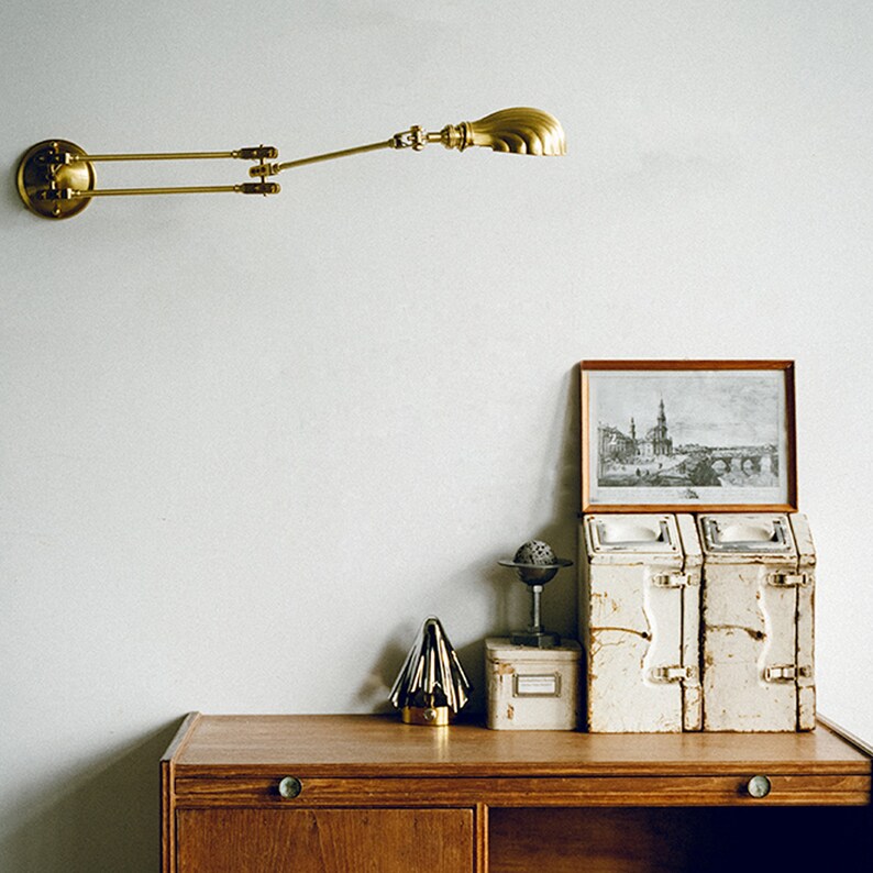 K&ouml;nnte beinhalten: Eine Wandleuchte aus Messing mit verstellbarem Arm ist an einer wei&szlig;en Wand befestigt. Darunter zeigt ein Holzschreibtisch ein gerahmtes Bild, zwei Vintage-Metallbeh&auml;lter, einen kleinen Globus und einen dekorativen goldenen Baum.