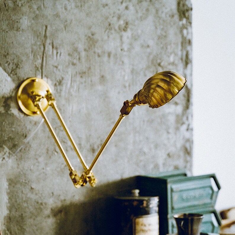 K&ouml;nnte beinhalten: Eine Wandleuchte aus Messing mit einem beweglichen Arm und einem gerippten Lampenschirm. Die Lampe ist an einer kreisf&ouml;rmigen Basis befestigt und an einer strukturierten grauen Wand montiert. Das Design der Lampe erinnert an industrielle Vintage-Beleuchtung.