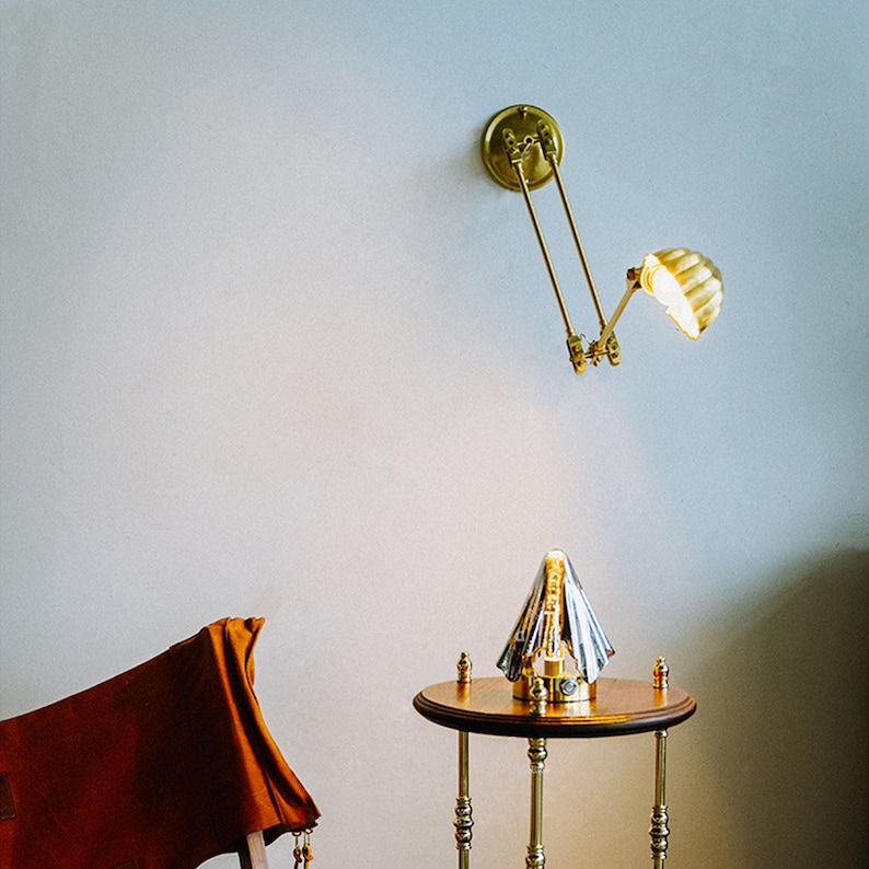 K&ouml;nnte beinhalten: Eine Wandlampe aus Messing mit verstellbarem Arm und schalenf&ouml;rmigem Schirm ist beleuchtet. Ein kleiner, runder Tisch mit Messingbeinen und einer Holzplatte tr&auml;gt eine dekorative Lampe. Ein brauner Stuhl befindet sich in der unteren linken Ecke.