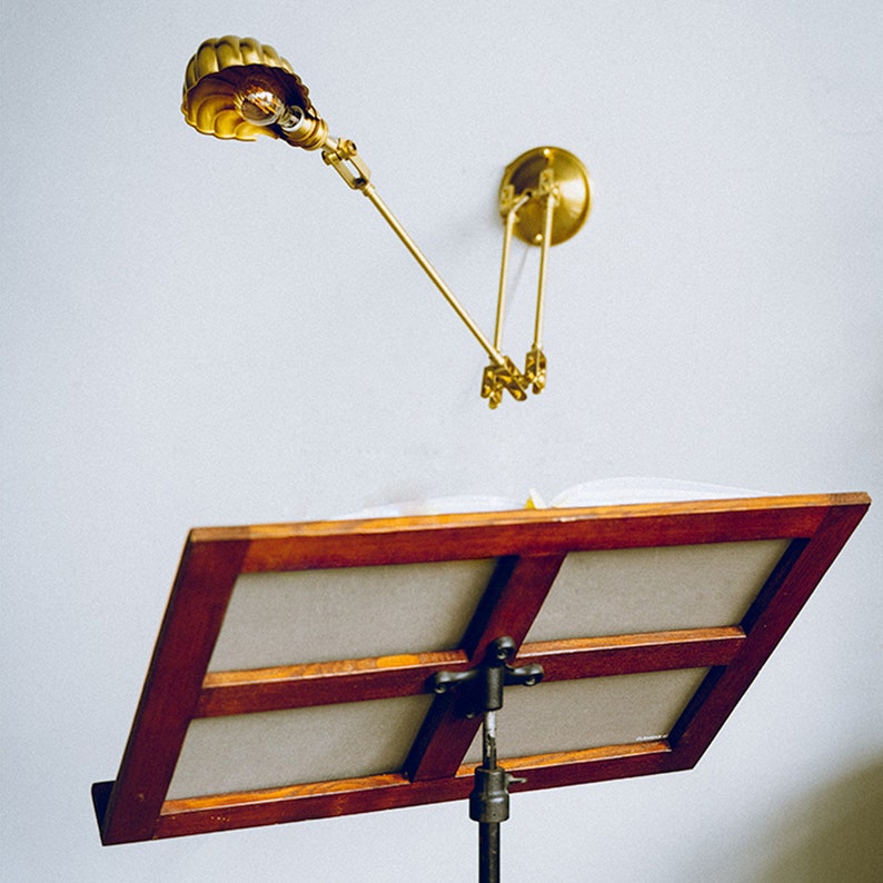 K&ouml;nnte beinhalten: Eine Wandleselampe aus Messing mit verstellbarem Arm und muschelf&ouml;rmigem Schirm beleuchtet einen h&ouml;lzernen Notenst&auml;nder. Der St&auml;nder h&auml;lt ein offenes Buch. Die Lampe ist an einer wei&szlig;en Wand befestigt.