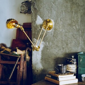 K&ouml;nnte beinhalten: Eine Wandleuchte aus Messing mit verstellbarem Arm und einem muschelf&ouml;rmigen Schirm. Die Lampe ist an einer strukturierten grauen Wand befestigt. Die Lampe ist eingeschaltet und strahlt ein warmes, gelbes Licht aus. Die Lampe ist Teil einer Wohnkultur-Kollektion.