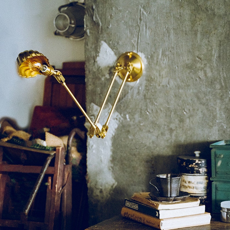 K&ouml;nnte beinhalten: Eine Wandleuchte aus Messing mit verstellbarem Arm und einem muschelf&ouml;rmigen Schirm. Die Lampe ist an einer strukturierten grauen Wand befestigt. Die Lampe ist eingeschaltet und strahlt ein warmes, gelbes Licht aus. Die Lampe ist Teil einer Wohnkultur-Kollektion.
