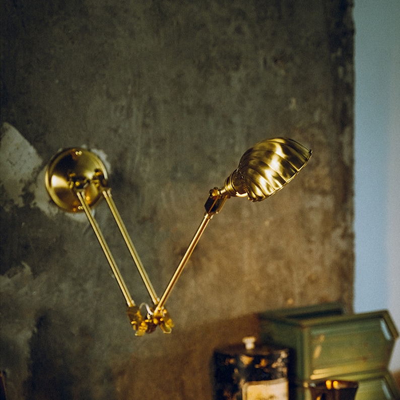 K&ouml;nnte beinhalten: Eine Wandleuchte aus Messing mit einem verstellbaren Arm und einem strukturierten, muschelf&ouml;rmigen Schirm. Die Lampe ist an einer grauen Wand montiert, und das Design erinnert an Vintage-Industriebeleuchtung.