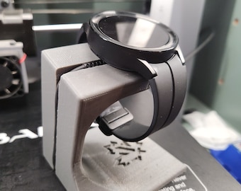 Puede incluir: Un soporte de impresión 3D para un reloj inteligente negro con una correa negra. El soporte tiene un pequeño diseño de copo de nieve en la base. El soporte está sobre una superficie negra con una etiqueta de advertencia sobre las temperaturas elevadas de la superficie.