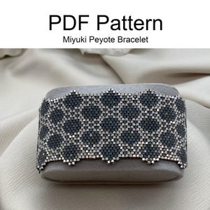 Può includere: Un braccialetto Miyuki peyote nero e argento con un motivo geometrico. Il braccialetto è esposto su un supporto grigio.