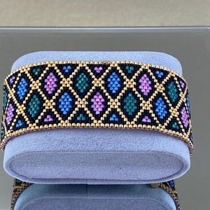 Op de afbeelding: Een kralenarmband met een geometrisch patroon van roze, blauwe, groene en gouden kralen op een zwarte achtergrond.