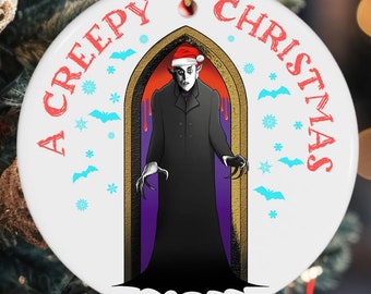 May include: A white ceramic Christmas ornament with a red and white "A Creepy Christmas" text. The ornament features a vampire wearing a Santa hat standing in a doorway with a purple and gold frame.