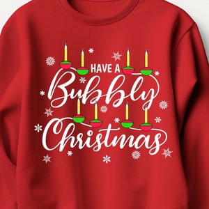 May include: Red Christmas sweatshirt with white text that says "Have a Bubbly Christmas". The text is surrounded by white snowflakes and a string of green and red Christmas lights.