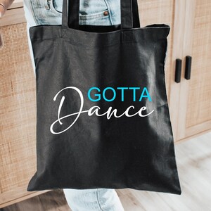 Op de afbeelding: Een zwarte tote bag met witte cursieve tekst die "Gotta Dance" in blauw zegt.