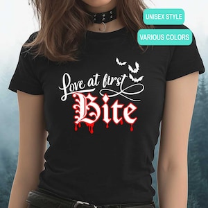 May include: A black short-sleeved t-shirt featuring the white text "Love at first" above the word "Bite" in a stylized red and white font with dripping red accents. Three small white bat silhouettes are positioned above the text.