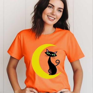 May include: A bright orange short-sleeved t-shirt featuring a graphic print of a black cat with green eyes sitting on a yellow crescent moon. The shirt has a crew neckline and is worn with blue denim jeans, showcasing a casual style.
