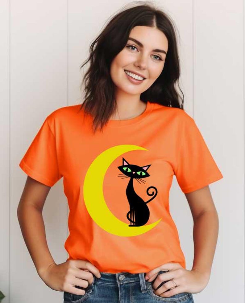 May include: A bright orange short-sleeved t-shirt featuring a graphic print of a black cat with green eyes sitting on a yellow crescent moon. The shirt has a crew neckline and is worn with blue denim jeans, showcasing a casual style.