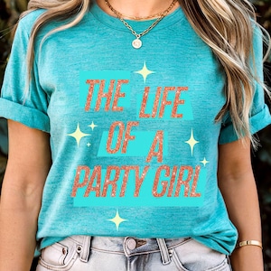May include: Teal short-sleeved t-shirt with the text "THE LIFE OF A PARTY GIRL" in glittery orange letters. The text is set against a light blue rectangular background, with yellow starburst graphics. The shirt is made of a soft material.