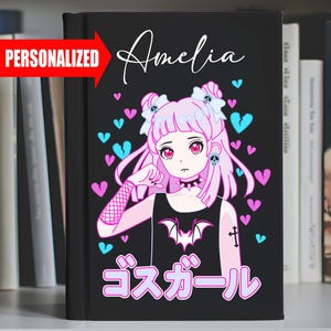 May include: A black notebook with a pink and blue anime girl illustration. The girl has pink hair, a black tank top with a bat design, and a cross tattoo on her arm. The text "Amelia" is written in white at the top of the notebook. The text "ゴスガール" is written in pink at the bottom of the notebook.