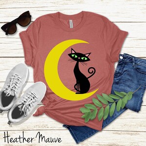 May include: A heather mauve short-sleeve t-shirt featuring a graphic of a black cat with green eyes sitting on a yellow crescent moon, accompanied by white trainers, dark blue denim jeans, and black sunglasses, with the text Heather Mauve.