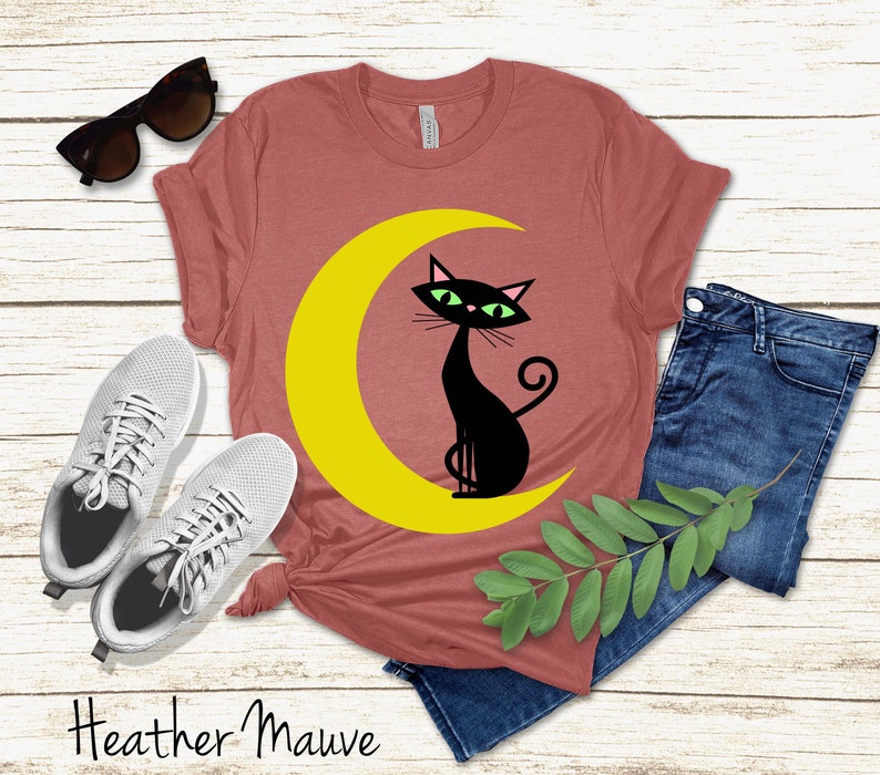 May include: A heather mauve short-sleeve t-shirt featuring a graphic of a black cat with green eyes sitting on a yellow crescent moon, accompanied by white trainers, dark blue denim jeans, and black sunglasses, with the text Heather Mauve.
