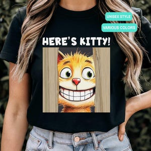 May include: Black t-shirt featuring a cartoon cat peeking through a wooden fence, with the text "HERE'S KITTY!". The cat has orange fur, green eyes, and a large, toothy grin. The shirt also has the text "UNISEX STYLE" and "VARIOUS COLORS".