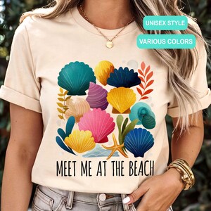 May include: Beige unisex t-shirt featuring a colorful seashell design with the text "MEET ME AT THE BEACH". The design includes various seashells in shades of blue, pink, yellow, and purple. The shirt is labeled "UNISEX STYLE" and "VARIOUS COLORS".