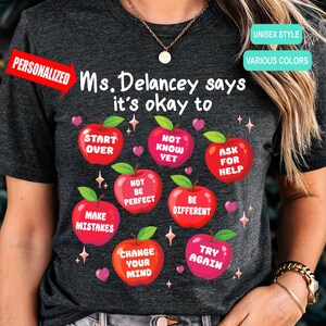 May include: A dark heather gray t-shirt featuring the text Ms. Delancey says it's okay to, surrounded by seven red apple illustrations. Each apple contains white text: Start Over, Not Know Yet, Ask For Help, Not Be Perfect, Be Different, Make Mistakes, Change Your Mind, and Try Again.