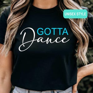 May include: A black short-sleeved t-shirt featuring the text "GOTTA" in bright blue and "Dance" in white script lettering. A small rectangular teal label in the upper right corner contains the white text "UNISEX STYLE".