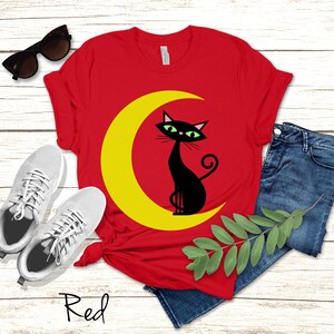 May include: A red t-shirt featuring a graphic of a black cat sitting on a yellow crescent moon. The shirt is displayed alongside a pair of white trainers, blue denim jeans, and black sunglasses. The word Red is written in black script below the shirt.