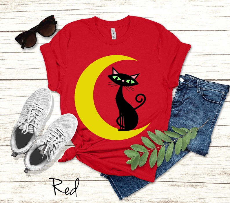 May include: A red t-shirt featuring a graphic of a black cat sitting on a yellow crescent moon. The shirt is displayed alongside a pair of white trainers, blue denim jeans, and black sunglasses. The word Red is written in black script below the shirt.