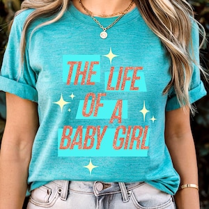 May include: A turquoise t-shirt with rolled-up sleeves. The shirt features the text "THE LIFE OF A BABY GIRL" in orange letters outlined in white, set against light blue rectangles. Gold starburst graphics are also present.