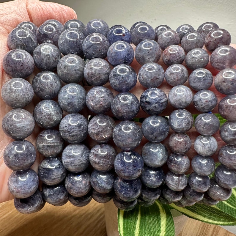 Pode incluir: Um fio de contas de pedras preciosas azul escuro e cinza. As contas s&atilde;o redondas e lisas, e t&ecirc;m um brilho ligeiramente iridescente.