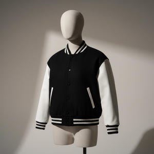 May include: A black and white varsity jacket displayed on a mannequin. The jacket features a black body with white sleeves. The collar, cuffs, and hem have white stripes. The jacket has a button-up front and two front pockets.