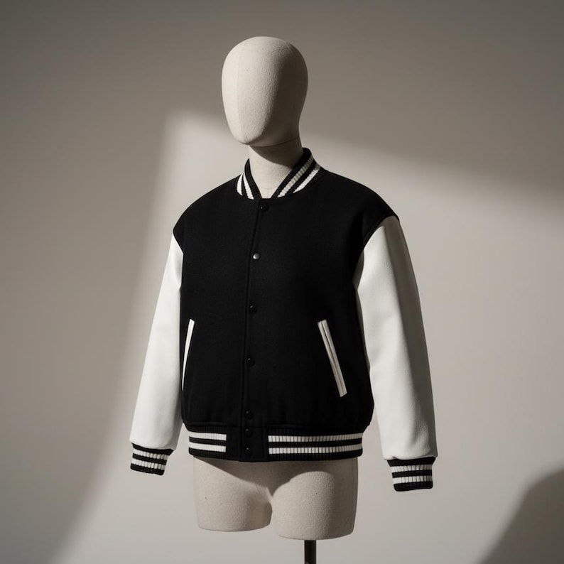 May include: A black and white varsity jacket displayed on a mannequin. The jacket features a black body with white sleeves. The collar, cuffs, and hem have white stripes. The jacket has a button-up front and two front pockets.