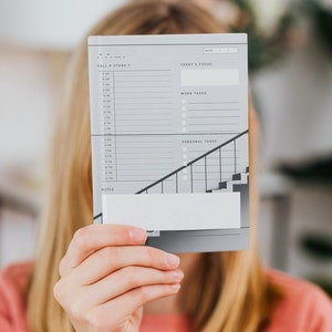 May include: A gray and white daily planner with a staircase design. The planner has sections for "Today's Focus", "Work Tasks", and "Personal Tasks". It also includes a section for notes and a time schedule from 5 AM to 12 PM.