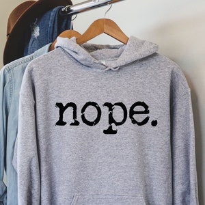 May include: A light gray hoodie with the word "nope." printed in black. The hoodie has a hood and a front pocket. It is hanging on a wooden hanger with other clothing items.