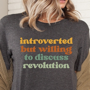 May include: A dark gray t-shirt with the text "introverted but willing to discuss revolution" in colorful retro lettering.