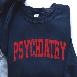 May include: A navy blue sweatshirt with the word "PSYCHIATRY" printed in red on the front. The sweatshirt is laid flat on a white surface with a pair of gray canvas shoes and blue jeans.