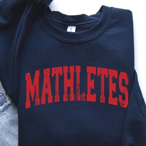 May include: A navy blue sweatshirt with the word "MATHLETES" printed in red on the front. The sweatshirt is laid out on a white surface with a pair of gray sneakers and a pair of blue jeans.
