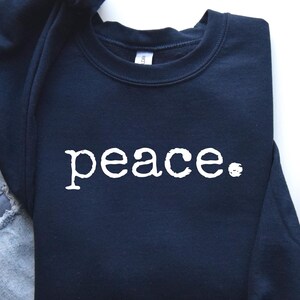 May include: A navy blue sweatshirt with the word "peace" printed in white. The sweatshirt is folded on a white surface next to a pair of light blue jeans, a brown mug with coffee, and gray sneakers.