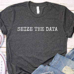 May include: A dark gray t-shirt with the text "SEIZE THE DATA" printed in white on the front. The shirt is laid flat on a white wooden surface with a pair of white sneakers and blue jeans.