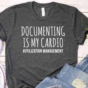 May include: A dark heather gray short-sleeve t-shirt featuring the white printed text "DOCUMENTING IS MY CARDIO" followed by "#UTILIZATION MANAGEMENT" in a smaller font. The shirt is displayed alongside white canvas sneakers and blue jeans.