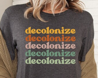 May include: A dark gray t-shirt with the word "decolonize" repeated five times in a rainbow of colors. The shirt is being worn by a person with long brown hair.