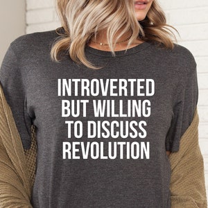 May include: A dark gray t-shirt with the text "Introverted but willing to discuss revolution" printed in white.