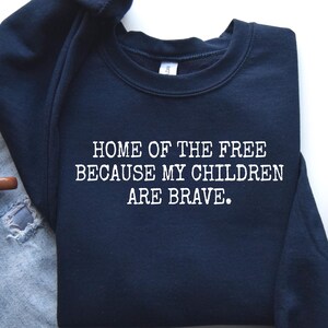 May include: Navy blue sweatshirt with the text "HOME OF THE FREE BECAUSE MY CHILDREN ARE BRAVE" in white. The sweatshirt is paired with light blue jeans, a cup of coffee, and gray sneakers.