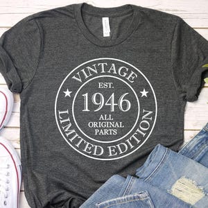May include: Dark gray t-shirt with a white circular graphic that reads "VINTAGE EST. 1946 ALL ORIGINAL PARTS LIMITED EDITION." The shirt is paired with blue jeans and white sneakers.