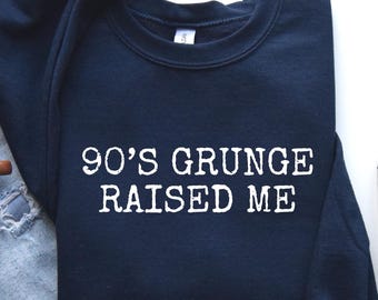 May include: A navy blue sweatshirt with the text "90'S GRUNGE RAISED ME" in white. The sweatshirt is paired with light blue jeans, a brown mug with coffee, and gray sneakers.