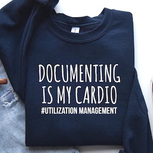 May include: A navy blue crewneck sweatshirt featuring the white text "DOCUMENTING IS MY CARDIO" and "#UTILIZATION MANAGEMENT" printed on the front, accompanied by light blue distressed denim jeans, a brown mug of coffee, and gray sneakers.
